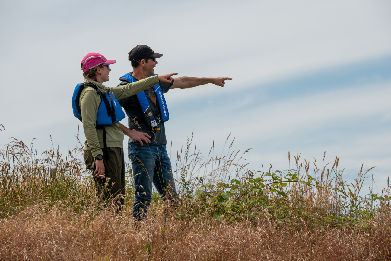 People standing in tall grass with lifejackets on pointing in the distance.